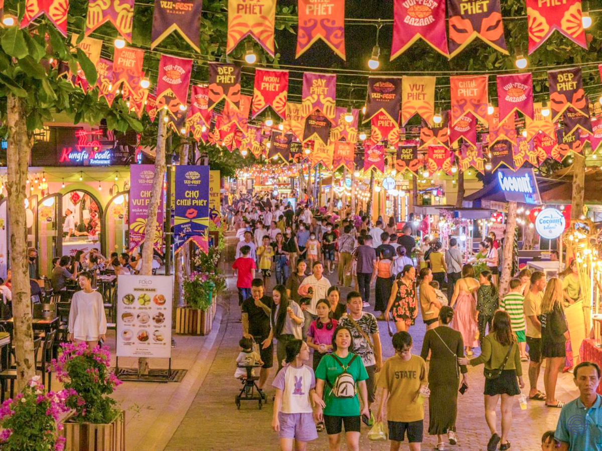 People stroll through a vibrant night market in Phu Quoc, lit by colorful banners and string lights.