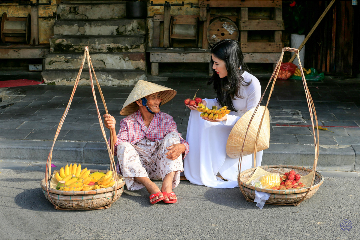 A woman in a white áo dài selects fresh fruit from a smiling vendor in Ho Chi Minh City.