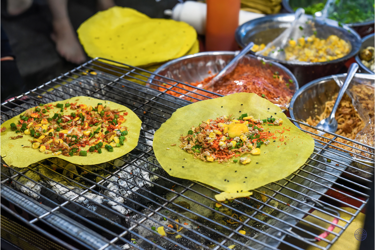 Delicious Vietnamese street food pancakes, topped with colorful ingredients, cooking on a grill in Ho Chi Minh City.