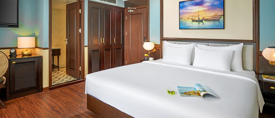 A luxurious cruise cabin featuring a comfortable white bed, elegant wooden decor, and a vibrant seascape painting.