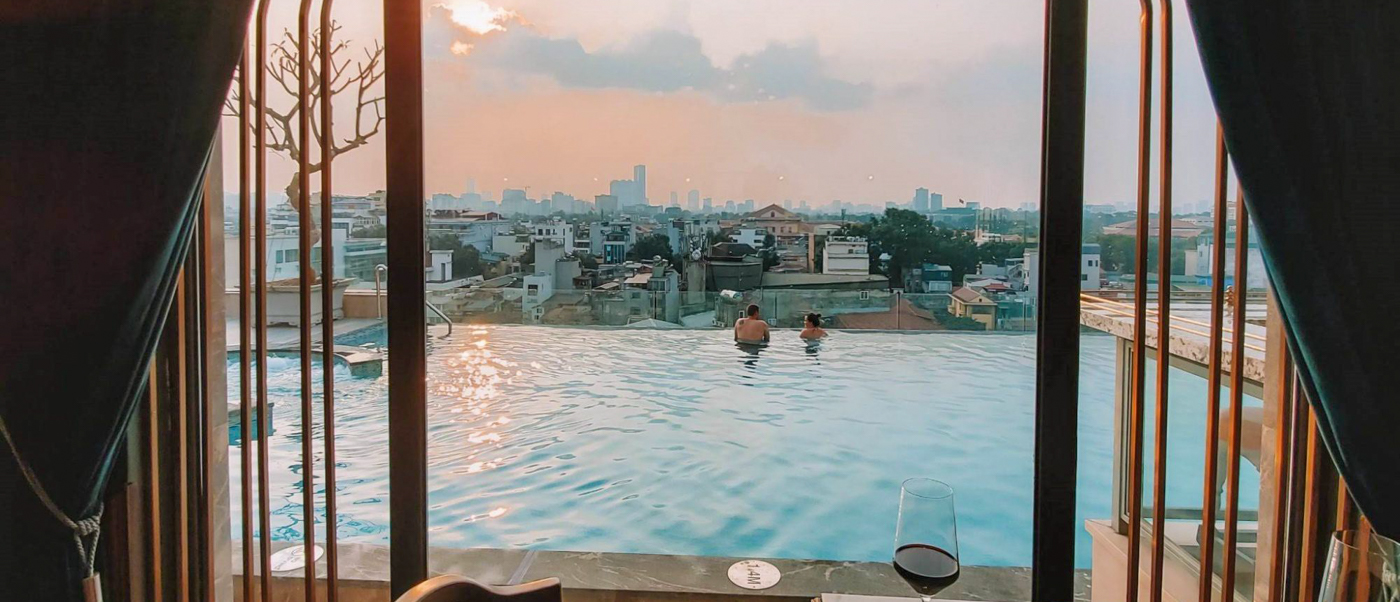 A couple relaxes in a rooftop infinity pool overlooking the Hanoi city skyline at sunset.
