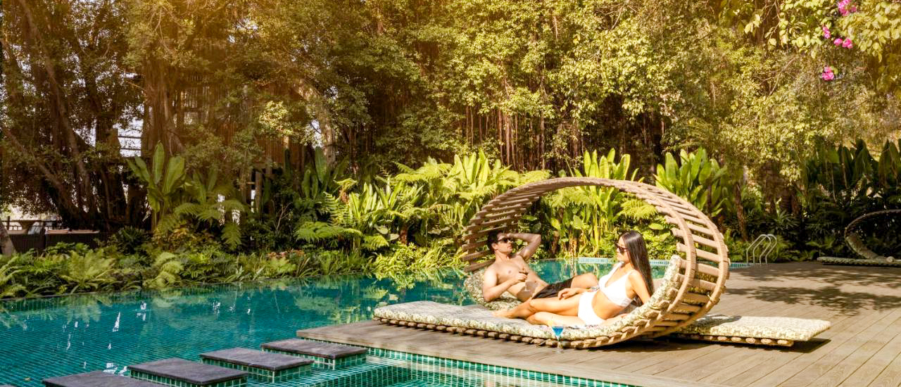 A couple relaxes on a unique circular lounge chair beside a sparkling pool and lush tropical plants.