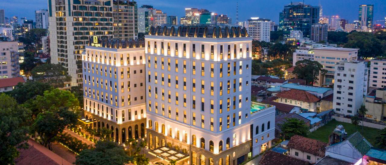 Elegant Mai House Saigon Hotel illuminated at dusk, towering over the vibrant Ho Chi Minh City skyline.