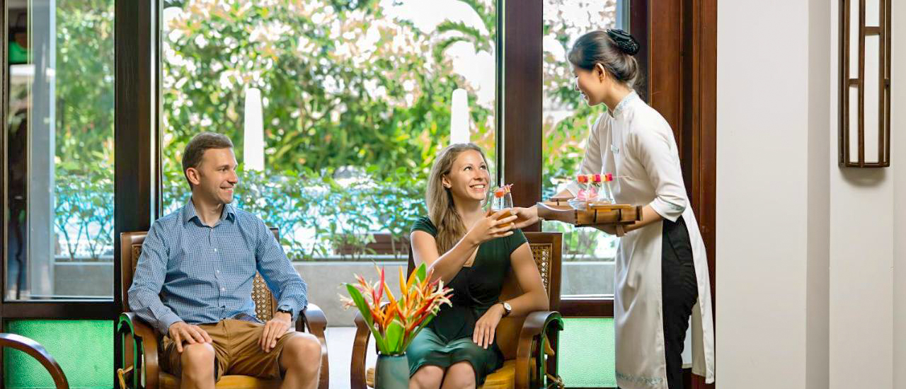 A smiling couple receives welcome drinks from a hotel staff member in Hoi An, Vietnam.