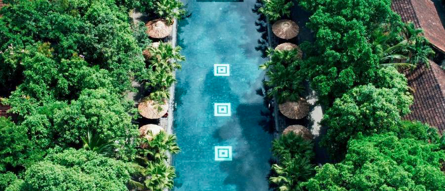 Overhead view of a tranquil blue pool bordered by lush green foliage and thatched cabanas in Hue.