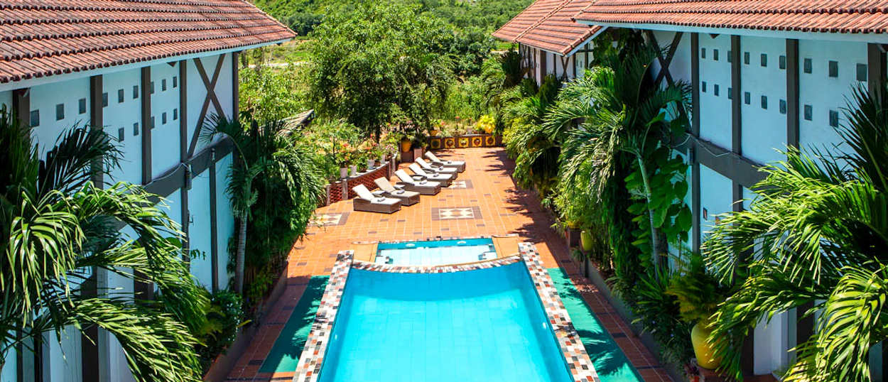 A serene resort swimming pool with sun loungers, nestled between buildings and lush tropical greenery.