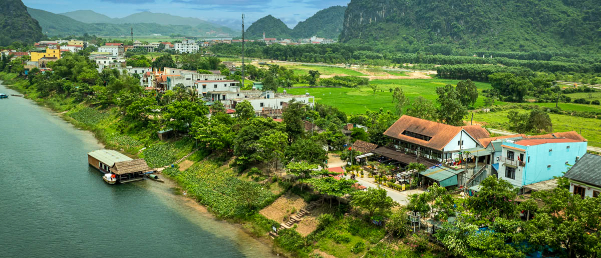 A peaceful river scene in Phong Nha, Vietnam, with a village nestled among green fields and karst mountains.