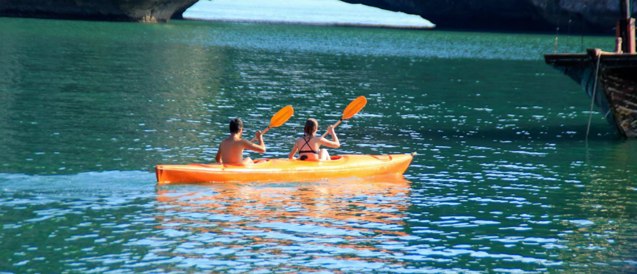 A couple paddles an orange kayak on the tranquil emerald waters of Halong Bay.