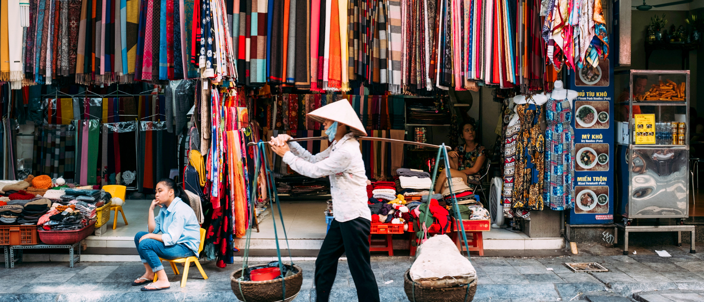 A street vendor in a conical hat carries baskets past a colorful fabric shop in Hanoi.