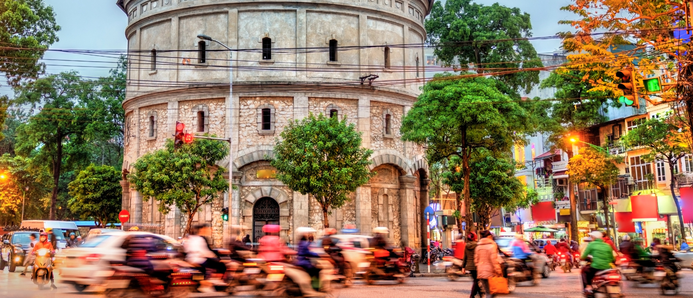 Lively evening street in Hanoi with many motorbikes and cars moving past a stone tower.