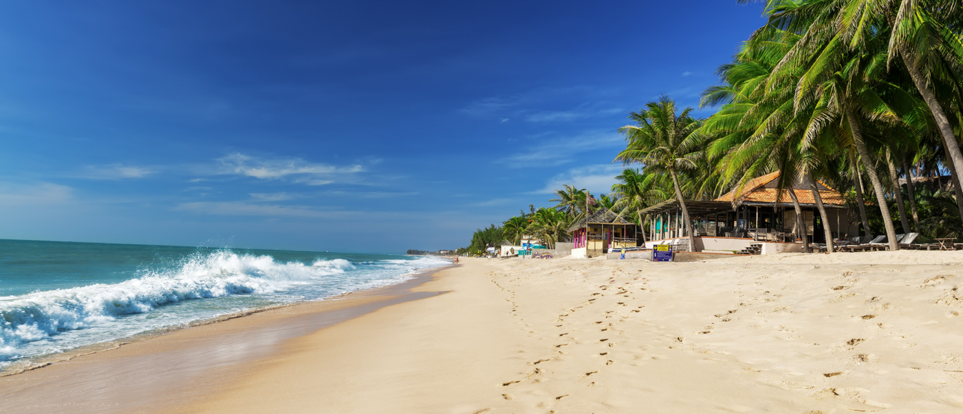 Waves lap a sunny sandy beach lined with palm trees and beach huts in Hoi An, Vietnam.