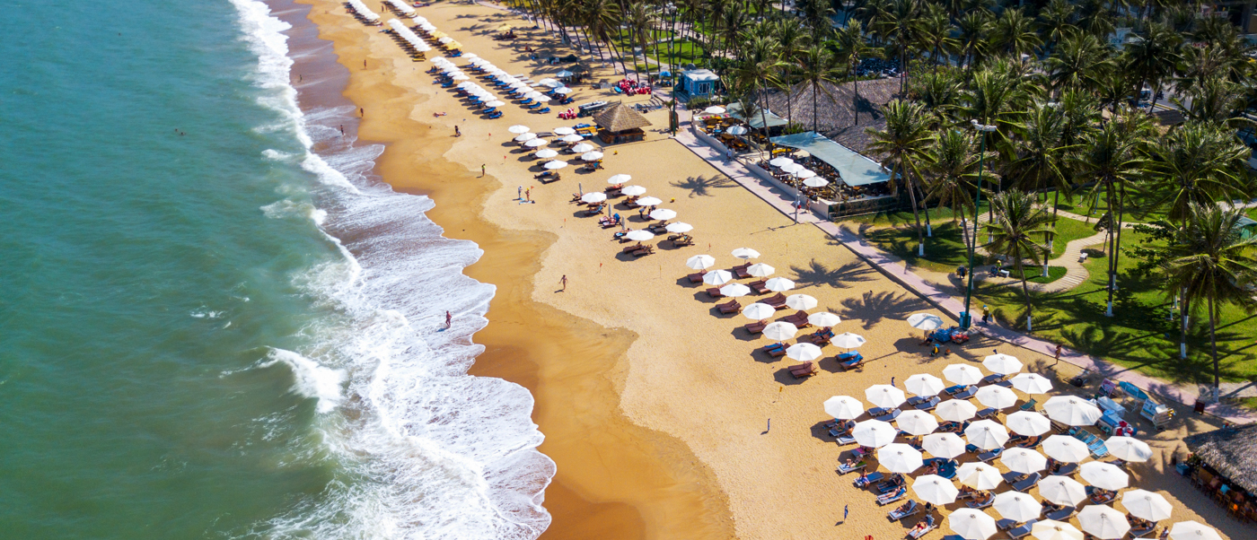 Aerial view of An Bang Beach in Hoi An, Vietnam, with rows of umbrellas, people, and palm trees.