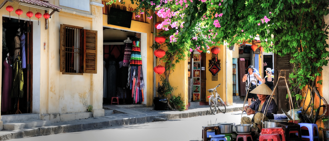 Hoi An street vendor in a conical hat prepares food under vibrant pink bougainvillea and red lanterns.