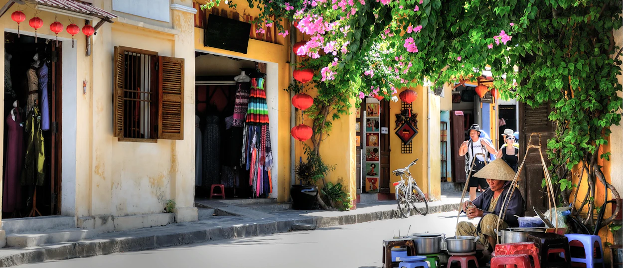Hoi An street vendor in a conical hat prepares food under vibrant pink bougainvillea and red lanterns.