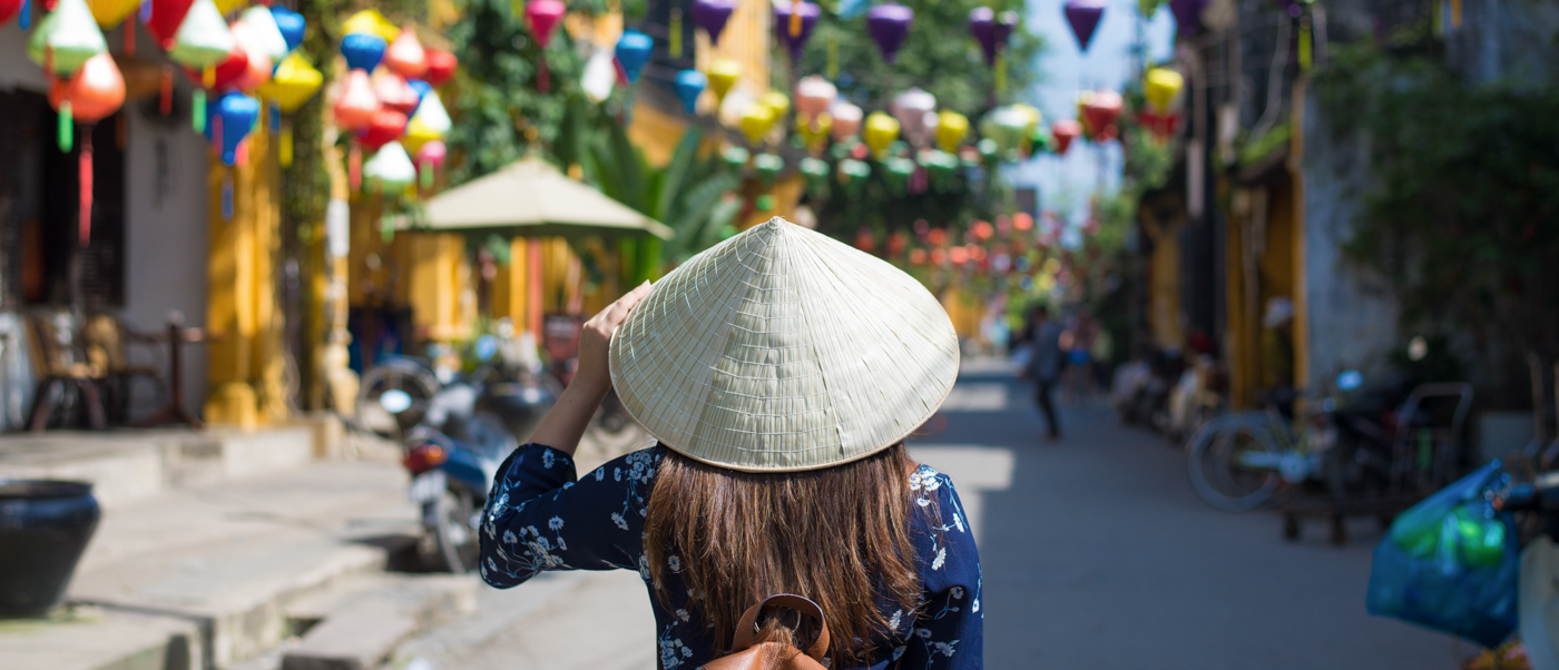 Person in conical hat walking down a vibrant street decorated with colorful lanterns in Hoi An.