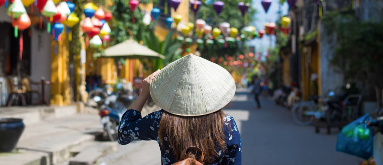 Person in conical hat walking down a vibrant street decorated with colorful lanterns in Hoi An.