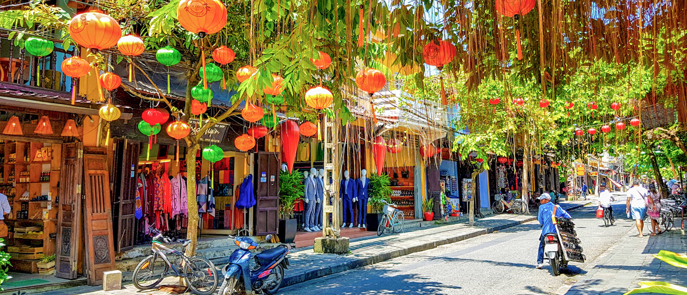 A charming street in Hoi An, Vietnam, with colorful lanterns, lush trees, and people walking.