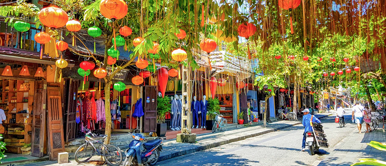A charming street in Hoi An, Vietnam, with colorful lanterns, lush trees, and people walking.