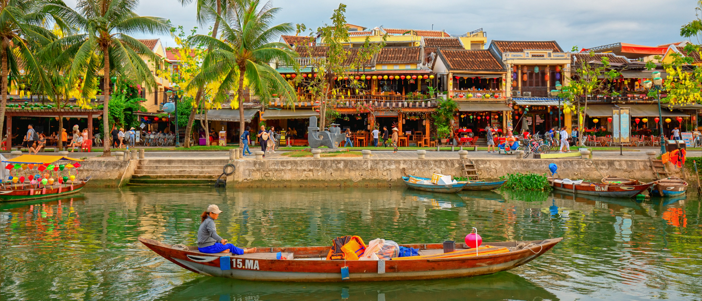 A person enjoys a bamboo boat ride past charming riverside buildings and palm trees in Hoi An.