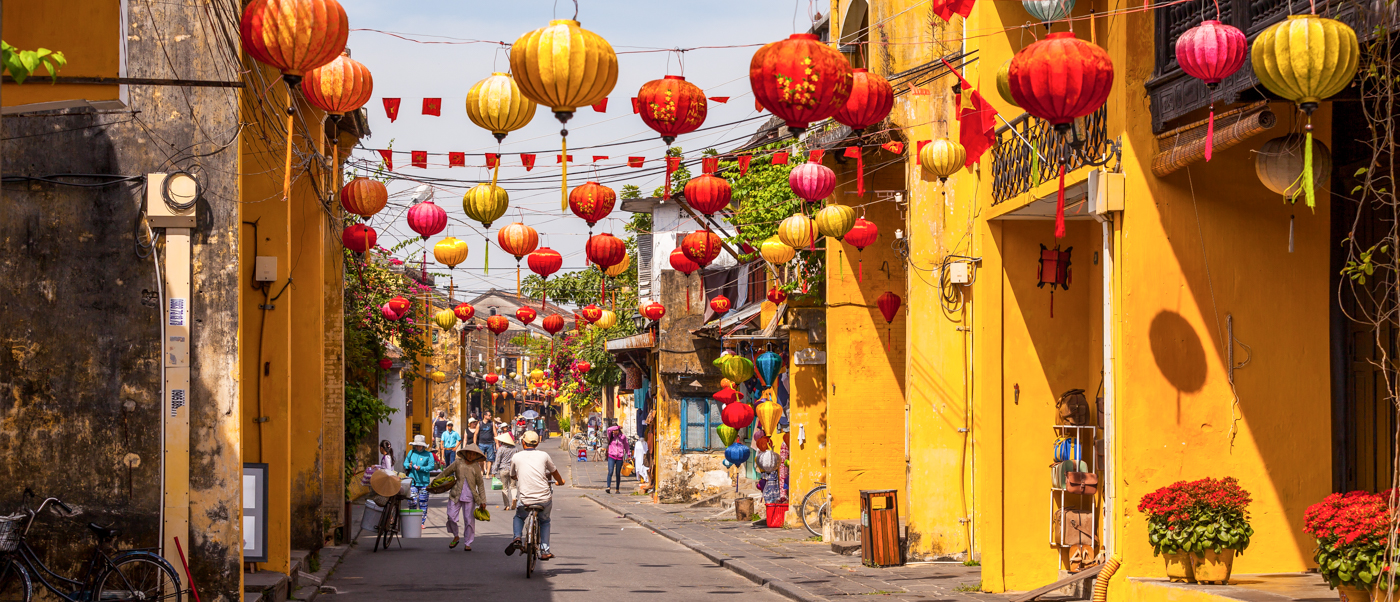 Colorful lanterns hang over a bustling street in Hoi An, lined with charming yellow buildings and people.
