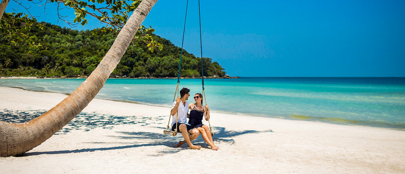 A happy couple sits on a swing under a palm tree on a beautiful white sand beach in Phu Quoc.