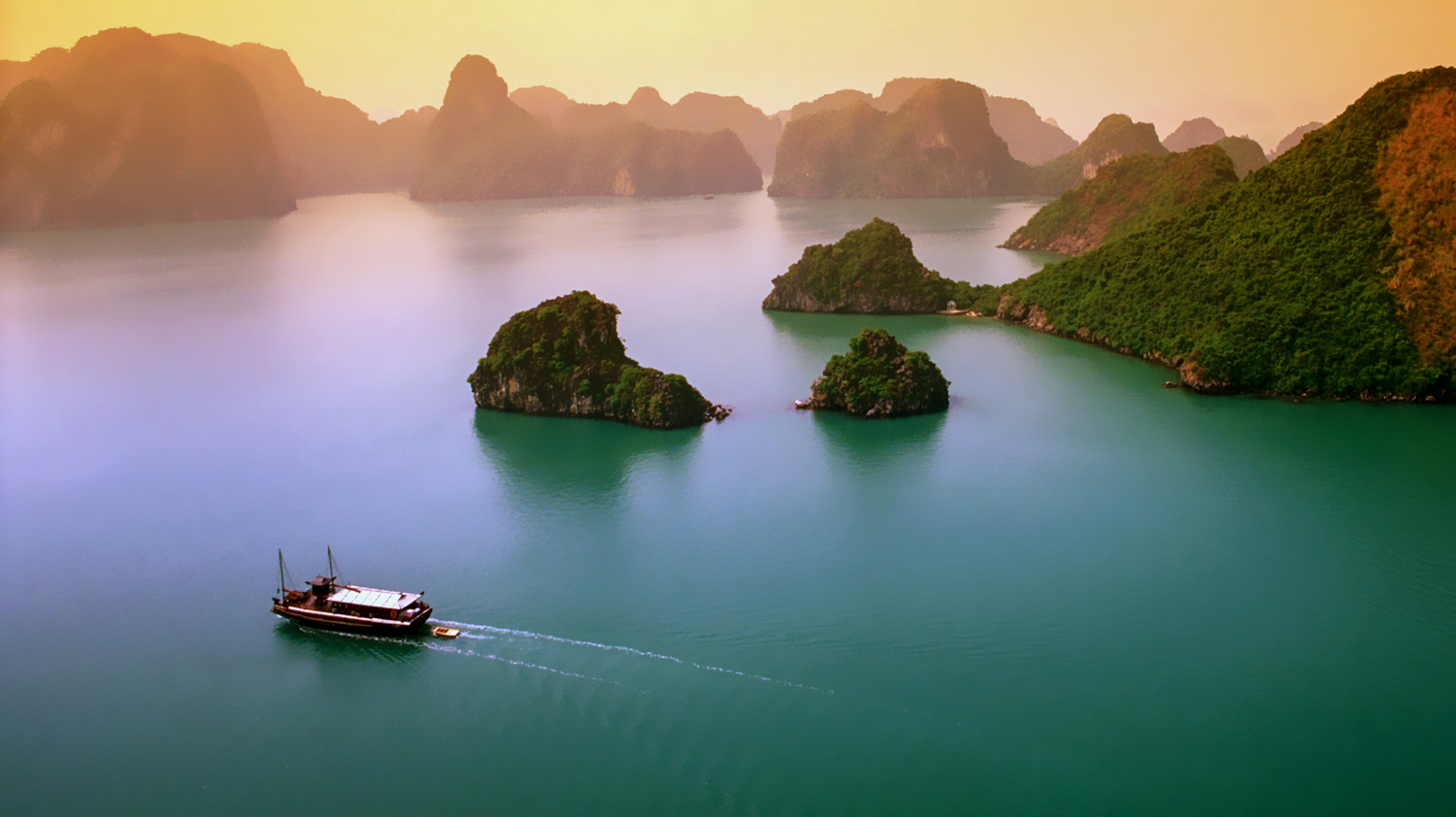A traditional boat sails on emerald waters surrounded by majestic limestone karsts at sunset in Halong Bay.