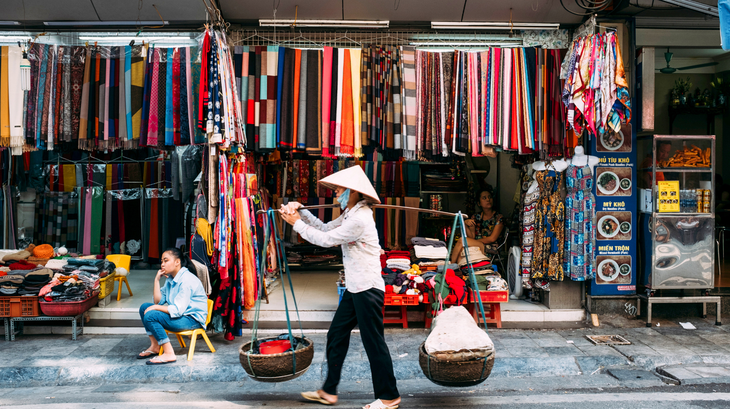 A vendor in a conical hat carries goods past vibrant textile stalls in a bustling Hanoi market.